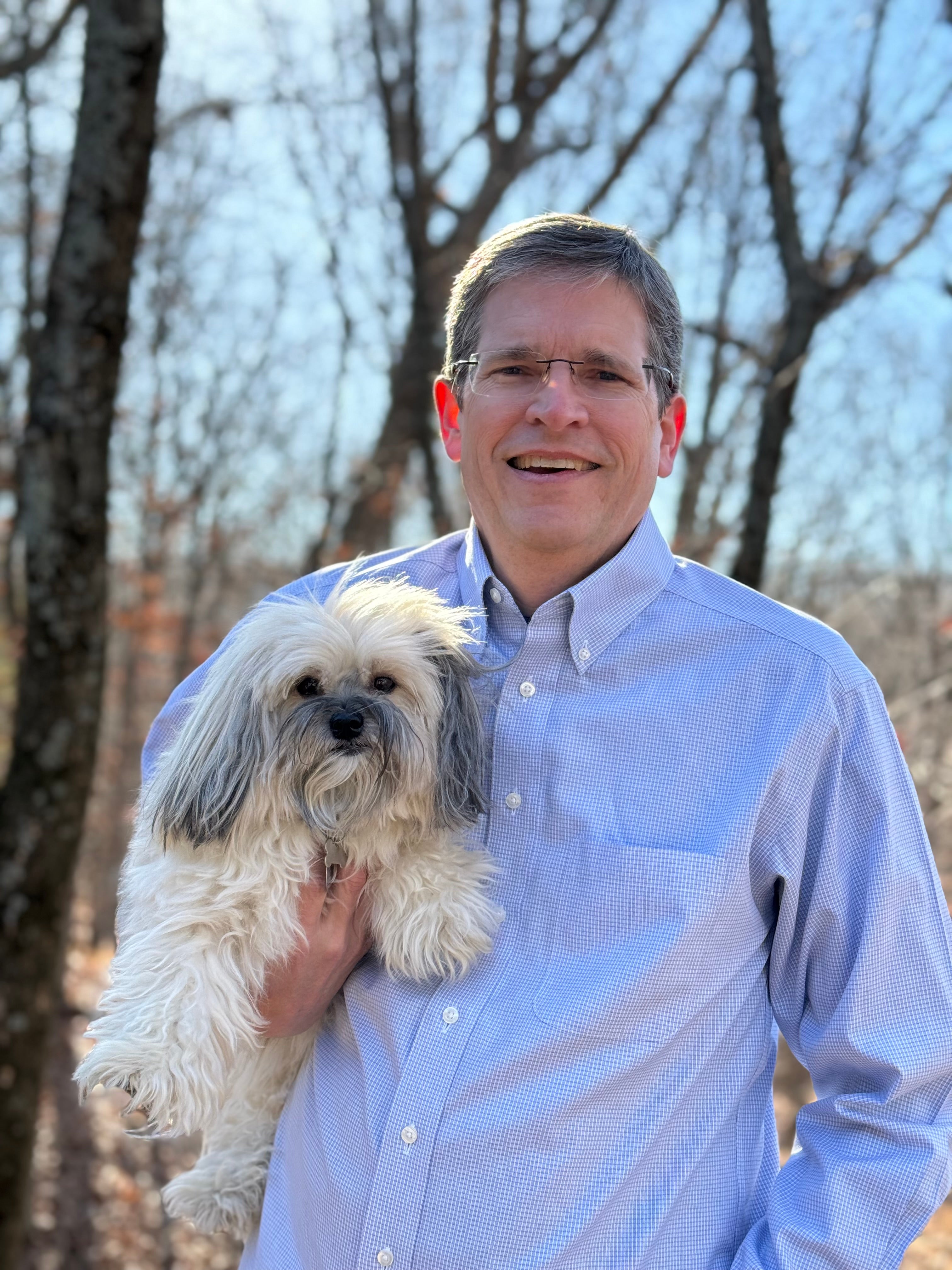 This image is of Bryan Monninger, Vice President of Vet Sales and Marketing at YuMOVE, with a small fluffy white dog in front of trees.