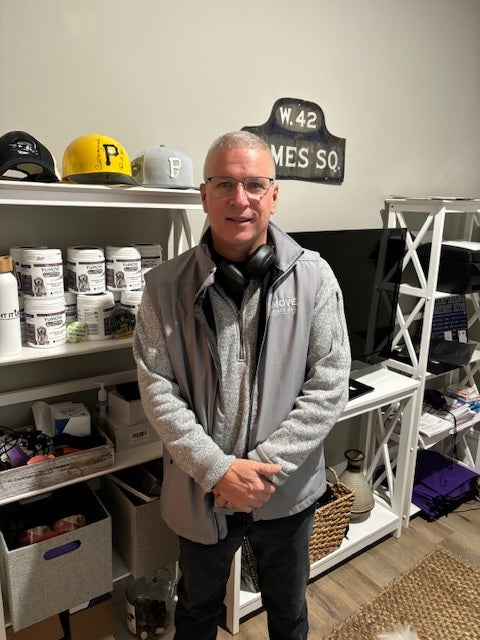 A man with glasses wearing a gray jacket and headphones around his neck, standing in a room with shelves containing various items, including hats and containers."