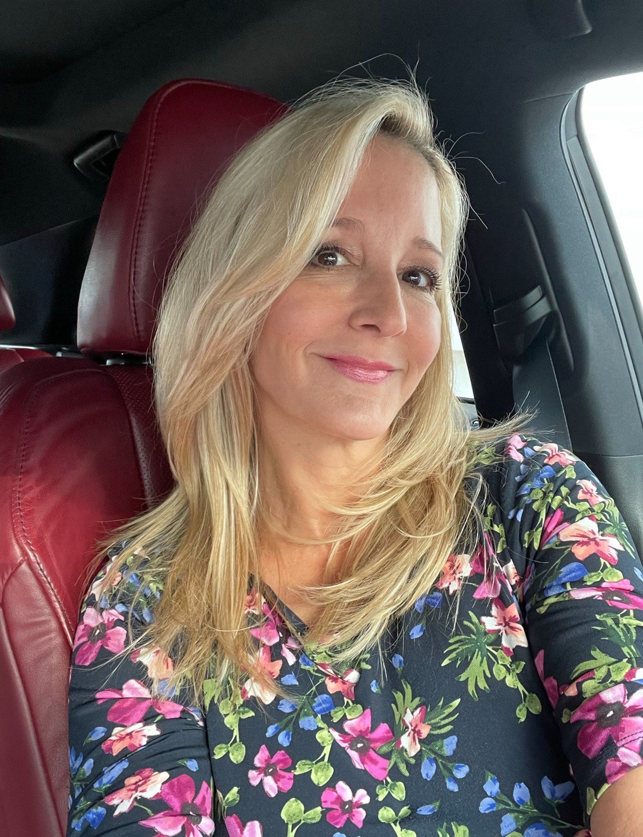A selfie of a woman with blonde hair, wearing a floral top, seated in a car with red leather seats.