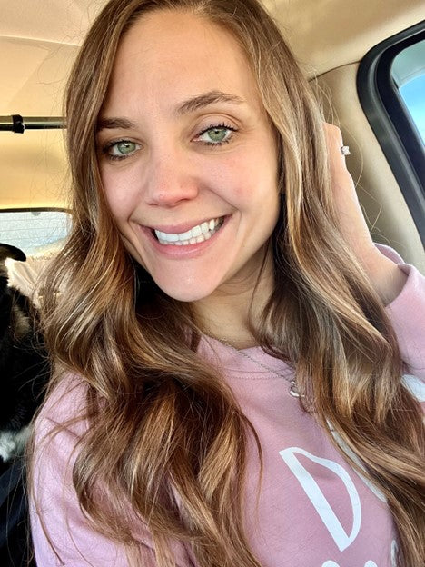 A woman with long brown hair and green eyes, wearing a pink hoodie, smiling in a car.