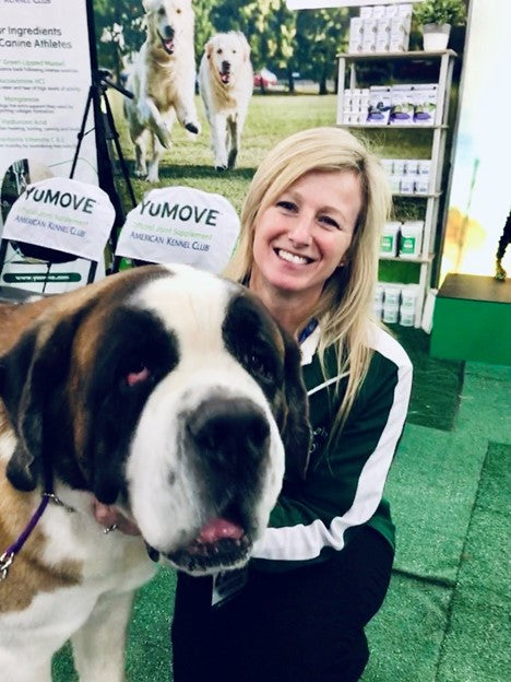 GPT A woman smiling at the camera with a large Saint Bernard dog on a leash, at a promotional event with banners for dog products in the background.